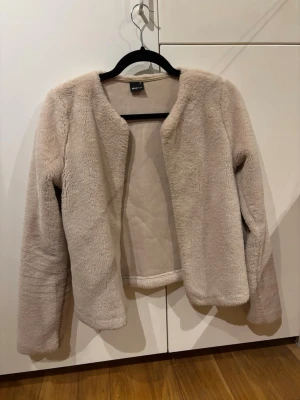 Beige teddyjacka från Only - Mysig beige teddyjacka från Gina Tricot med öppen front och rundad halsringning. Jackan har långa ärmar och är gjord i ett fluffigt, mjukt material som ger en avslappnad och trendig look. Perfekt för att hålla värmen under kyliga dagar.