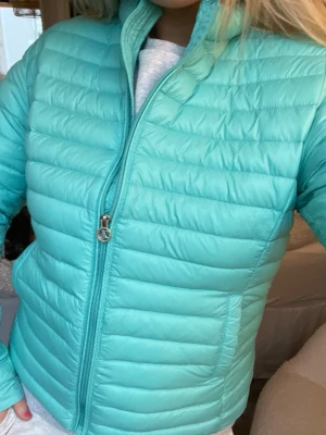 Turkos pufferjacka med dragkedja - Snygg turkos pufferjacka med quiltad design och hög krage. Jackan har dragkedja framtill och två sidofickor. Perfekt för kalla dagar och ger en sportig vibe. Materialet är lätt och isolerande, vilket håller dig varm under vintern.