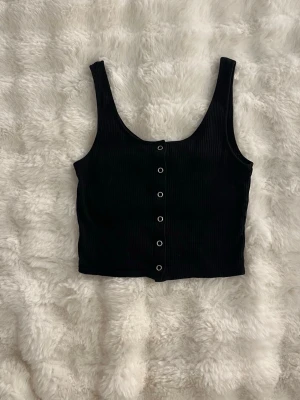 Svart ribbad linnetopp med knappar - Snygg svart ribbad linnetopp med knappar framtill. Croppad modell med breda axelband och rundad halsringning. Perfekt att styla med höga jeans eller kjol för en trendig look. Inga defekter förekommer! Bild med plagg på kan skickas vid begäran. Postar inom 1-2 dagar😇 Ligger även ute på min vinted:ellyssellout