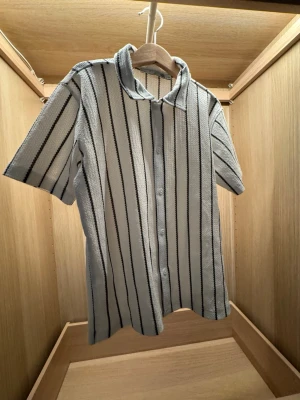 Randig kortärmad skjorta - Säljer en vit skjorta med svarta och grå vertikala (randiga) ränder. Skjortan har klassisk krage, knappar framtill och korta ärmar. Materialet känns som bomull eller polyester och ger en clean och fräsch vibe.