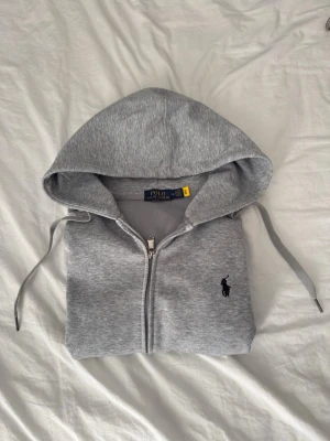 Grå zip hoodie från Polo Ralph Lauren - Snygg grå hoodie med dragkedja från Polo Ralph Lauren. Klassisk design med huva ärmar med ribbade muddar sopm ger en clean look. sopm ger en clean look. Liten svart logga broderad på bröstet. Tillverkad i mjukt bomullsmaterial och har fickor framtill. Storlek m och passar mig som är 180