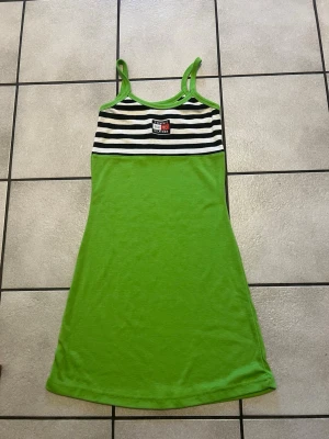 Grön randig klänning Tommy Hilfiger - Snygg grön klänning från Tommy Hilfiger med smala axelband och svartvit randigt mönster upptill. Klänningen har en broderad logga på bröstet och är gjord i mjukt bomullstyg. Perfekt för sommarens alla tillfällen!