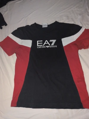 Svart EA7 Emporio Armani t-shirt - Snygg svart t-shirt från EA7 Emporio Armani med vita och röda detaljer på ärmarna. Stor logga framtill i vitt. Klassisk passform och rund hals. Perfekt för dig som gillar streetwear och märkeskläder. Säljer pga för liten