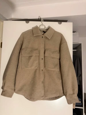 Beige teddy overshirt med fickor - Overshirt i beige teddy-material med två stora bröstfickor och knappar framtill. Rak passform, klassisk krage och långa ärmar med knapp vid ärmslut. Perfekt för lager på lager och ger en avslappnad men trendig look.