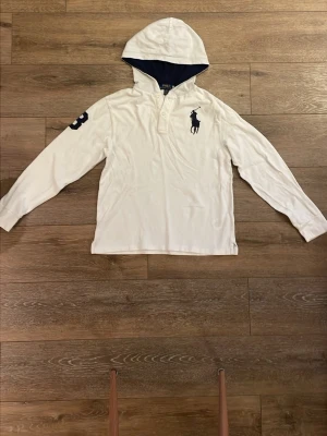 Vit hoodie från Ralph Lauren - Vit hoodie från Polo Ralph Lauren med mörkblå insida i huvan och klassisk logga broderad på bröstet. Långärmad modell med knappslå vid halsen och en stor siffra på ena ärmen. Tillverkad i mjukt bomullsmaterial. Priset går att ändra.