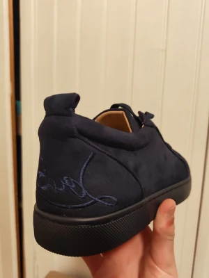 Mörkblå sneakers från Christian Louboutin - Snygga mörkblå sneakers från Christian Louboutin med klassisk röd sula och broderad logga på hälen. Skorna har en låg profil och är tillverkade i mocka med svarta snören. Perfekta för dig som vill sticka ut med exklusiva detaljer.