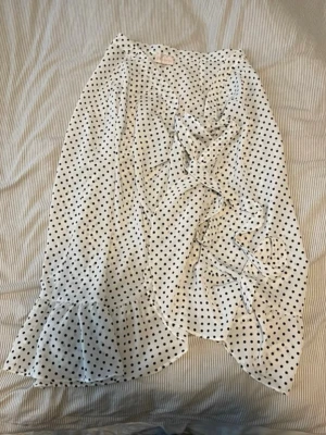 Vit prickig omlottkjol med volang - Säljer en vit omlottkjol med svarta prickar och volangkant. Kjolen har knytband i midjan.