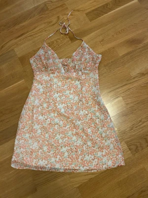  Ny Blommig kort klänning med knyt - Söt kort klänning med blommigt mönster i ljusa toner av rosa, vit och beige. Klänningen har smala axelband med knyt i nacken och v-ringad urringning. Perfekt för varma dagar och somriga vibes.