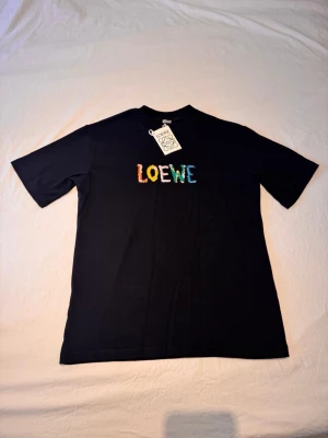 Svart Loewe t-shirt med färgglad logga - Snygg svart t-shirt från Loewe med broderad logga i flera färger på bröstet. Klassisk rund hals och korta ärmar. Tillverkad i mjuk bomull som känns skön mot huden. Perfekt för dig som vill ha en stilren men ändå unik look. Säljs då den aldrig kommit till användning. 