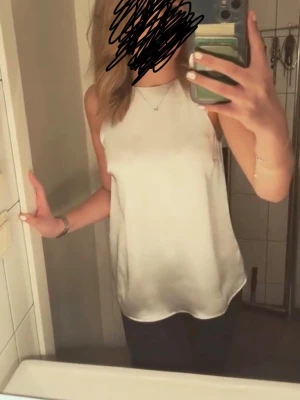 Champagne halterneck topp i satin - Snygg champagne linnetopp i satin med ärmlös design och rund halsringning. Toppen har en slät och glansig yta som ger en lyxig känsla och passar perfekt till jeans eller kjol. Enkel och stilren modell som är lätt att matcha.