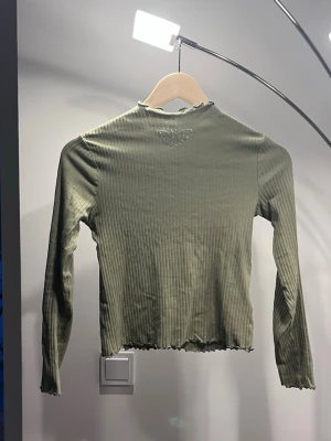 Olivgrön ribbad topp med fjärilsdetalj - Säljer en olivgrön ribbad långärmad topp med en glittrig fjäril i strass på ryggen. Toppen har vågig kant vid hals och nederkant för en extra söt touch. Perfekt att styla med jeans eller kjol.