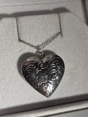 Silverfärgat hjärthalsband medaljong med mönster i rostfritt stål  - Säljer ett silverfärgat halsband med ett hjärtformat medaljonghänge. Hänget har vackra, ingraverade blommönster och kan öppnas för att lägga in ett foto eller minne. Kedjan är tunn och elegant, och hela halsbandet har en klassisk och romantisk stil. Kedja 45 cm i rostfritt stål och hänge ca 3 cm i rostfritt stål/metall.
