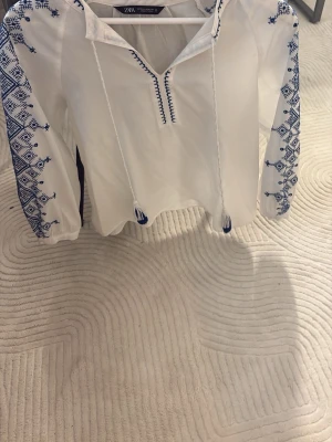 Vit blus med blå brodyr från Zara - Vit blus från Zara med blå broderade detaljer på ärmar och axlar. Blusen har långa ärmar, snörning med tofsar vid halsen. Pris går att diskutera vid snabb affär