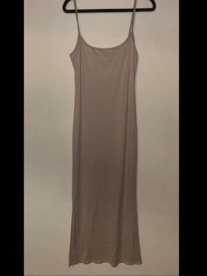 Beige slipklänning med smala axelband - En enkel och stilren beige slipklänning med tunna axelband och rak passform. Klänningen är hellång och har en minimalistisk design som passar perfekt till lager på lager eller sommarens varma dagar. Materialet är mjukt och den är inte see-through 