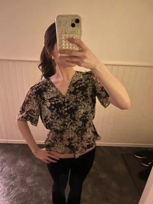 Blommig blus med  volanger - Säljer en svart blommig blus med v-ringning och korta ärmar. Blusen har volangdetalj i midjan och ett avslappnat fall. Perfekt för dig som gillar romantiska mönster och en feminin look. Den är köpt utomlands på en marknad så jag vet inte storlek på den. Jag har vanligtvis 34,36 passar mig perfekt 