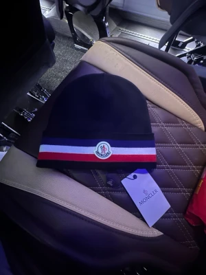 Moncler mössa med röd och vit rand - Snygg mörkblå Moncler mössa med breda ränder i vitt och rött längst ner samt Moncler-logga framtill. Mössan är stickad och har en klassisk form som passar perfekt för kyliga dagar. Perfekt accessoar för dig som gillar stilrena detaljer och exklusiva märken. Lappen finns kvar 