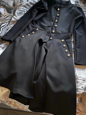 Svart rock med guldknappar från Crown - Stilren svart rock från Crown med dekorativa guldknappar i dubbla rader doorway framtill och på sidorna. Rocken har en markerad midja och klassisk krage. Materialet är slätt och ger ett elegant intryck. Perfekt för dig som vill sticka ut med sopren look.