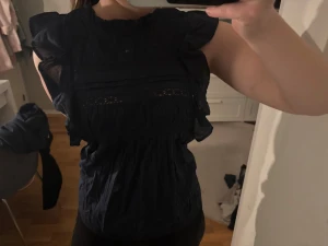 Mörkblå volangblus med spets - Säljer en mörkblå sopranblus från Holly & Whyte med volangdetal path längs ärmhålen och spetsinslag framtill. Blusen är ärmlös och har en hög halsringning. Materialet känns lätt och luftigt, perfekt för en trendig och avslappnad look. Storlek S, väldigt väldigt få gånger använd🌸