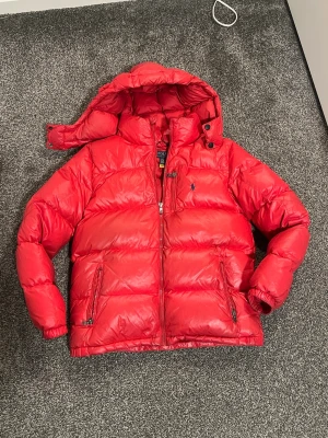Röd Ralph Lauren dunjacka (glansig) – strl 14–16 – avtagbar luva - Säljer en röd glansig Ralph Lauren jacka i storlek 14–16. Köpt på KidsBrandStore. Väldigt snygg och varm jacka.  • Avtagbar luva • Flera fickor med dragkedja • Skön och varm  Skick: Bra skick men använd. Finns en liten lagning på ärmen, klädhängaren i nacken har gått isär och några små missfärgningar, men de syns knappt.  Nypris runt 3000 kr.  Skriv om du vill ha fler bilder eller info. 