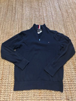 Mörkblå stickad tröja Tommy Hilfiger - Mörkblå stickad tröja från Tommy Hilfiger med halv dragkedja och ståkrage. Tröjan har diskret logga på bröstet och ribbade muddar vid ärmslut och nederkant. Röd, vit och blå randig detalj i kragen. Det står storlek L men jag skulle säga att det är storlek M