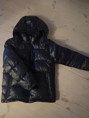 Svart dunjacka från Polo Ralph Lauren - Säljer : en svart dunjacka från Polo Ralph Lauren med.. huva och dragkedja framtill. Jackan har två sidofickor med dragkedja och en liten röd logga på bröstet. Perfekt för kalla vinterdagar och riktigt skön att bära.