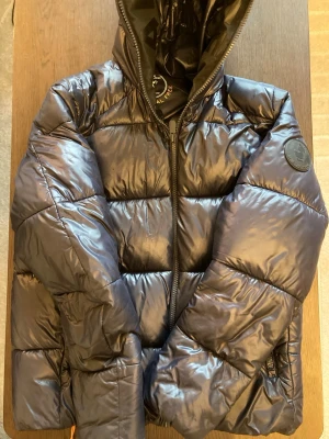 Marinblå dunjacka från Save The Duck - Snygg marinblå dunjacka från Save The Duck med glansig finish och vadderad design. Jackan har huva, dragkedja framtill och en rund logotyp på ärmen. Perfekt för kalla vinterdagar och riktigt trendig look. Den är i generellt väldigt bra skick och håller värmen bra. Ända att anmärka på är den lilla skadan bak på ryggen som du ser på bild 5 där jag råka stöta i något vasst men inget som påverkar och syns knappt heller.