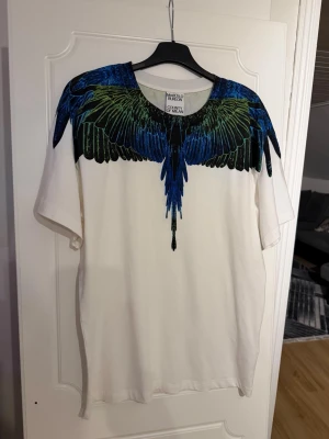 Marcelo Burlon t shirt  - Säljer en vit t-shirt från Marcelo Burlon med ett unikt tryck av blå och gröna vingar över axlar och bröst. T-shirten har rund halsringning och korta ärmar. Perfekt för dig som vill sticka ut med en cool design.