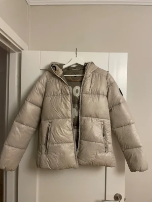 Beige pufferjacka från Save The Duck - Snygg beige pufferjacka från Save The Duck med huva och dragkedja framtill. Jackan är 100% animal free och har två praktiska fickor med dragkedja. Perfekt för kalla vinterdagar och har en modern, oversized look.