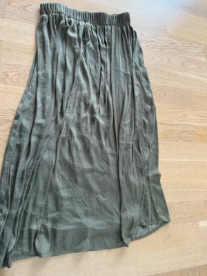 Olivgrön långkjol med resårmidja - Säljer en olivgrön långkjol med avslappnad passform och resår i midjan. Kjolen har ett tunt, lätt tyg som ger ett snyggt fall och passar perfekt till både sneakers och sandaler. Enkel och stilren design utan extra detaljer.