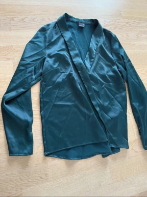 Mörkgrön omlottblus i satin - Stilren mörkgrön omlottblus i glansigt satinmaterial med djup v-ringning och långa ärmar. Perfekt för att lyfta vilken outfit som helst med sin eleganta look och mjuka känsla. Snygg detalj med omlott framtill.