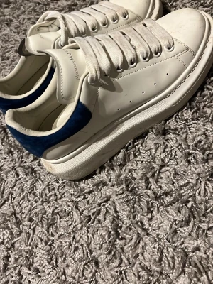 Alexander McQueen vita sneakers - Stilrena vita sneakers från Alexander McQueen med chunky sula och blå detalj på hälen. Skorna har perforerade sidor, breda vita skosnören och logga på plösen. Tillverkade i läder med en modern och clean look. Pris kan diskuteras 