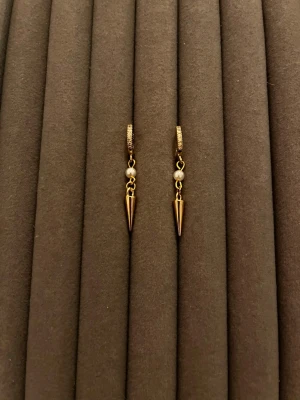 Guldiga örhängen med pärldetalj - Snygga örhängen i guldfärgad metall med små vita pärlor och spetsiga konformade hängen. Örhängena har en modern design med en liten pärldetalj som ger en cool kontrast till det guldiga metallet. Perfekta för att lyfta din stil.
