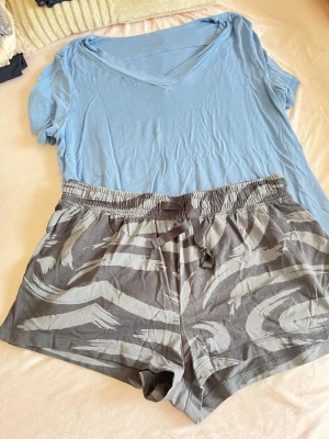 Blå pyjamas med mönstrade shorts - Mysig pyjamas med ljusblå t-shirt i mjukt material och v-ringning. Tillhörande shorts i grått med abstrakt mönster i ljusare toner och elastisk midja med dragsko. Perfekt för chillkvällar hemma.