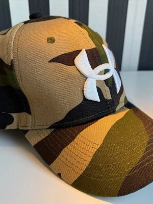 Camo keps från Under Armour - Säljer en snygg keps från Under Armour med camouflagemönster i grönt, brunt och beige. Stor vit logga broderad framtill. Tillverkad i 60% polyester och 40% bomull för skön passform. Perfekt för dig som gillar streetwear och sportig stil.