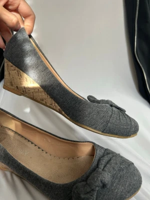 Grå ballerinaskor med kilklack - Snygga grå ballerinaskor med kilklack i kork och dekorativ rosett framtill. Skorna har en mjuk textilovandel och innersula i beige. Perfekta för dig som vill ha en bekväm men ändå stilren look.