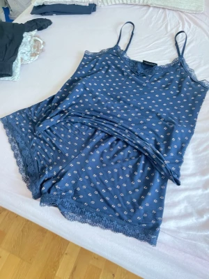 Blå pyjamas-set med spetsdetaljer - Mysigt blått pyjamas-set med linne och shorts. Setet har tunna axelband, små vita blommönster och fina spetsdetaljer vid halsringning och benslut. Perfekt för chillkvällar hemma. Materialet känns mjukt och luftigt.
