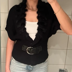 Svart kofta - Snygg svart kofta med korta ärmar, dekorativ fuskpäls längs kragen och ett brett midjebälte med stor metallspänne. Passar XS-M, jag har vanligtvis S. 55% ull, 45%bomull. Fattas två metallringar på bältet, dock syns inte detta när den är knäppt. 