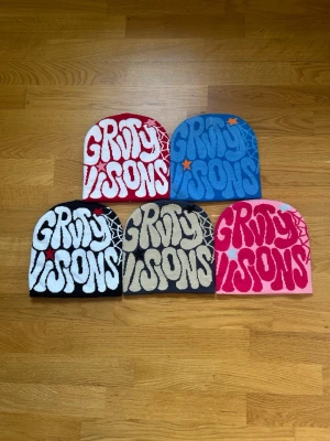 Groovy Visions mössa idx5 färgern - Fem stickade mössor>  från Groovy Visions med grafiskt tryck framtill och spindelnätsdetalj bak. Finns i röttöd, blått, svart, grått och rosa. Materialet är mjukt och stretchigt, perfekt för dig som vill sticka ut med cool streetwear.