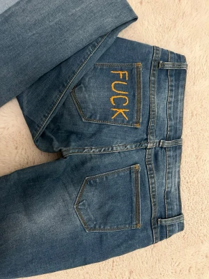 Lågmidjade jeans - Unika blå jeans med utsvängda ben och slitningar. Ena bakfickan har gul brodyr med texten 'FUCK' för en edgy look. Jeansen är med lågmidja