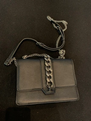 Svart väska  - Snygg svart hand still väska i konstläder med silverfärgad kedjedetalj på locket och sopig axelrem i både kedja och läder. Väskan har magnetlås och en stilren, rektangulär form som passar till många olika looks. Perfekt för dig som gillar en edgy och modern stil.