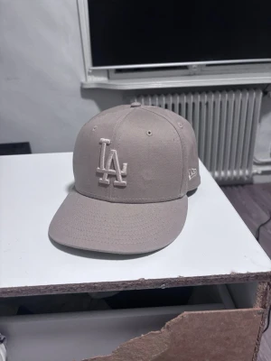 Beige LA-keps från New Era - Snygg beige keps från New Era med LA-logga broderad framtill. Klassisk modell med böjd skärm och broderade ventilationshål. Perfekt accessoar för en avslappnad streetwear-look.