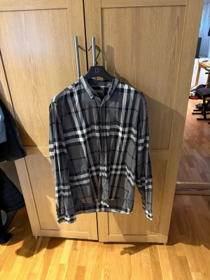 Grå burberry skjorta med  - Snygg burberry skjorta med stort rutigt mönster i vitt och svart. Skjortan har klassisk krage, långa ärmar och knäppning framtill. Perfekt för en avslappnad men ändå stilren look.