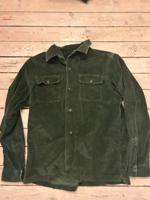 Gröna manchesterovershirt Polo Ralph Lauren - Mörkgröna manchesterbyxor från Polo Ralph Lauren med klassisk rak passform. Byxorna har två stora bröstfickor med lock och knappar, samt tydlig krage. Tillverkade i mjuk manchester för en retro och avslappnad look.