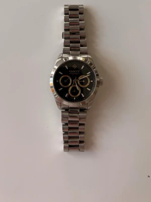 Silverfärgad GANT klocka med klockbox. - Stilren klocka från GANT med silverfärgad länk och svart urtavla. Urtavlan har flera små visare och detaljer i guld och silver. Klockan har en klassisk rund form och är tillverkad i metall, vilket ger en modern och tidlös look.