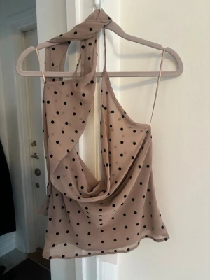 Beige prickig linnetopp med scarfdetalj - Trendig beige linnetopp med svarta prickar och tunna axelband. Toppen har en snygg scarfdetalj som kan knytas runt halsen och ett draperat fall framtill. Perfekt för dig som vill sticka ut med en unik och stilren look.