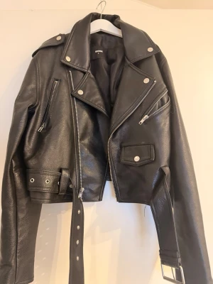 Svart skinnjacka från BikBok - Snygg svart skinnjacka från BikBok i klassisk bikerstil. Jackan har flera dragkedjor, axelklaffar, bred krage och ett bälte i midjan. Perfekt för dig som vill ha en edgy look med coola detaljer och tidlös design.