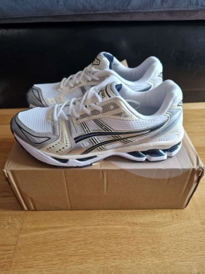Asics Gel-Kayano 14 vit/silver - Snygga Asics Gel-Kayano 14 sneakers i vitt och silver med svarta detaljer. Ovandelen är i mesh och syntet för bra andningsförmåga och komfort. Klassisk retrodesign med tydliga Asics-loggor på sidorna och GEL-dämpning i sulan.