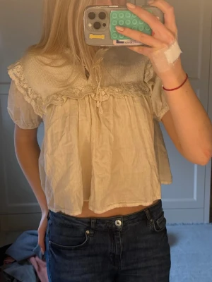 Beige blus med spets och volang - Söt beige blus med stor krage, spetsdetaljer och volangkant. Blusen har korta ärmar och knytband framtill vid halsen. Luftig och flowy passform, perfekt för en romantisk look.