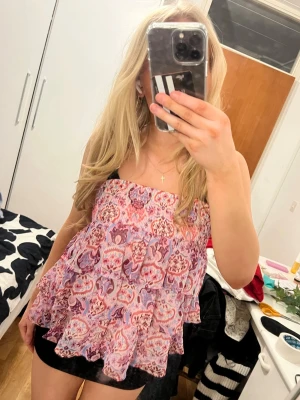 Blommig volangig bandeau topp - Säljer en söt bandeau topp med blommigt mönster i rosa och lila toner. Toppen har volanger och är axelbandslös, perfekt för sommarens varma dagar. Luftig och flowy modell som ger en avslappnad vibe.
