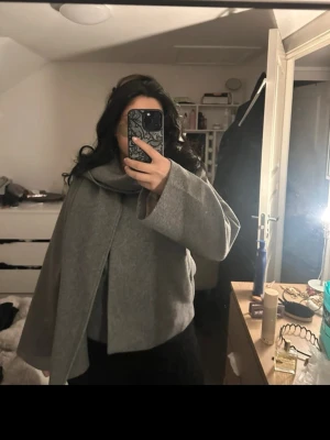 Grå cape-kappa med bred krage - Säljer en stilren grå cape-kappa med bred krage och vida ärmar. Kappan har en modern, boxig passform och är tillverkad i ett mjukt material som ger en elegant look. Perfekt för dig som vill ha något trendigt och bekvämt till hösten.